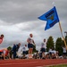 A group of military personnel are conducting a physical fitness test or training exercise on an outdoor running track. A blue unit guidon flag marked '142 MXG / 142 AMXS' is prominently displayed in the foreground, indicating this is a U.S. Air National Guard unit event. Participants are performing various exercises including push-ups and running, while two individuals in orange safety vests appear to be officiating or monitoring the event.