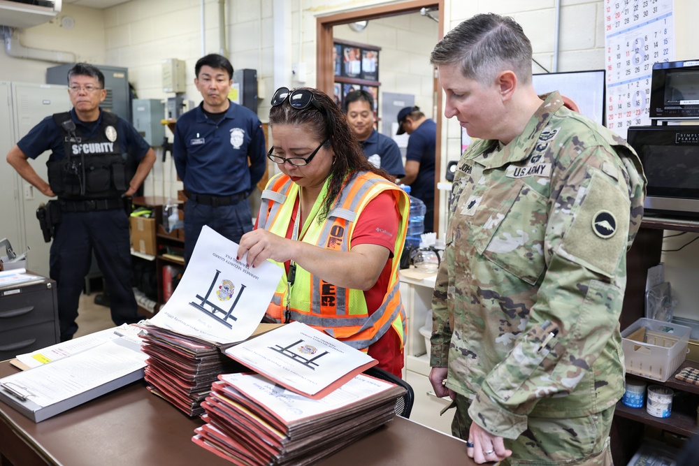 A U.S. Army soldier in OCP camouflage uniform reviews documents with a civilian worker wearing a high-visibility safety vest in what appears to be an operations or administrative office. Several security personnel in dark uniforms are visible in the background. The setting suggests an official coordination or inspection activity, possibly related to emergency management or facility operations.