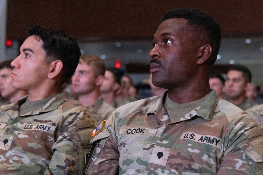 A group of U.S. Army soldiers in Operational Camouflage Pattern (OCP) uniforms are seated in formation, appearing to attend a briefing, ceremony, or presentation in a large indoor venue. The foreground soldier's name tape reads 'COOK' and displays a Specialist rank insignia. Multiple rows of soldiers are visible in the background, suggesting a large-scale military gathering.