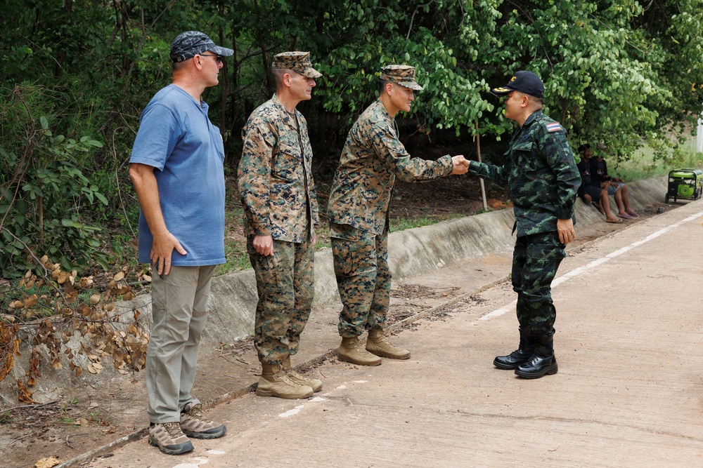 The image shows a meeting between military personnel on a paved outdoor path surrounded by trees. Two individuals in US Marine Corps MARPAT camouflage uniforms are present, one of whom is shaking hands with a person in a different camouflage uniform, likely Thai military based on the distinctive pattern and black cap with insignia. A civilian in a blue t-shirt and cap stands nearby observing the interaction.