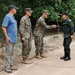 The image shows a meeting between military personnel on a paved outdoor path surrounded by trees. Two individuals in US Marine Corps MARPAT camouflage uniforms are present, one of whom is shaking hands with a person in a different camouflage uniform, likely Thai military based on the distinctive pattern and black cap with insignia. A civilian in a blue t-shirt and cap stands nearby observing the interaction.