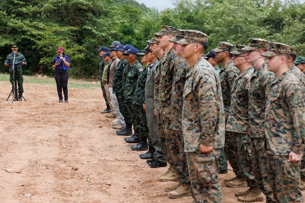 A military formation ceremony showing personnel from at least two different armed forces standing in a line on a dirt field. A speaker at a microphone stand addresses the assembled troops, with a civilian interpreter or aide standing nearby. The setting appears to be an outdoor training or ceremonial area surrounded by dense vegetation.