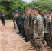A military formation ceremony showing personnel from at least two different armed forces standing in a line on a dirt field. A speaker at a microphone stand addresses the assembled troops, with a civilian interpreter or aide standing nearby. The setting appears to be an outdoor training or ceremonial area surrounded by dense vegetation.