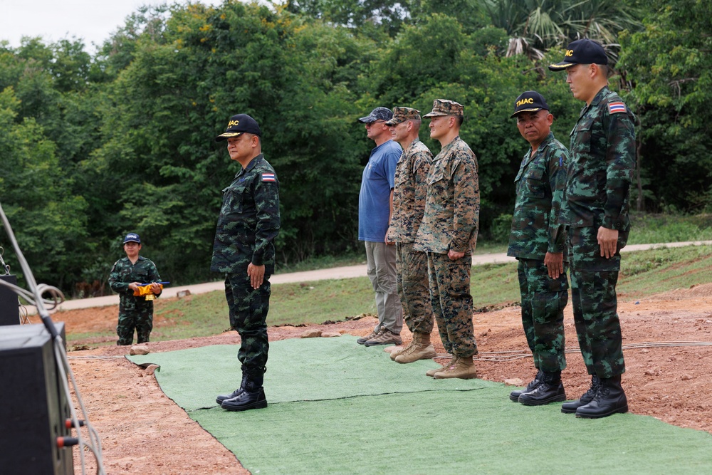 A group of military personnel are standing at attention on a green mat over red soil in an outdoor setting. The group includes individuals wearing Thai military camouflage uniforms with 'TMAC' caps bearing Thai flag patches, US Marine Corps MARPAT uniforms, and one civilian in casual clothing. The scene appears to be a formal military ceremony or inspection, likely a joint multinational exercise.