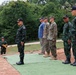 A group of military personnel are standing at attention on a green mat over red soil in an outdoor setting. The group includes individuals wearing Thai military camouflage uniforms with 'TMAC' caps bearing Thai flag patches, US Marine Corps MARPAT uniforms, and one civilian in casual clothing. The scene appears to be a formal military ceremony or inspection, likely a joint multinational exercise.