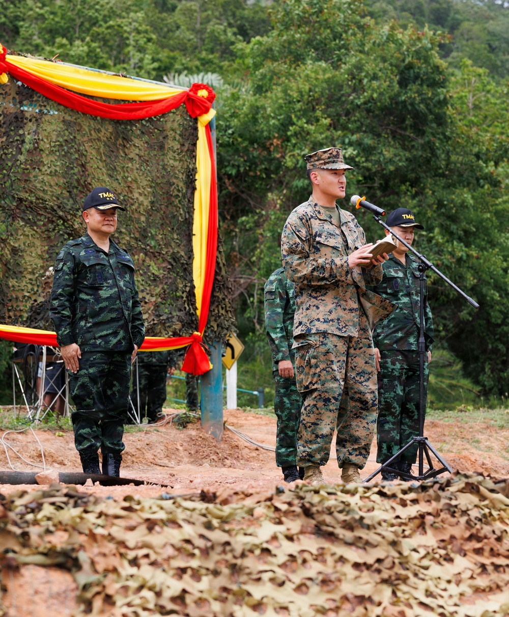 A military ceremony or briefing is taking place outdoors, featuring personnel in at least two distinct camouflage uniforms — one in US Marine Corps MARPAT digital camouflage and others in a darker woodland-pattern camouflage. The central figure, wearing USMC uniform and cover, is speaking at a microphone stand while holding a document. The setting is decorated with red and yellow draped fabric against a camouflage netting backdrop, suggesting a joint military exercise or formal event.