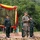 U.S. Marines and Thailand Mine Action Center participate in Deployed for Training Bilateral EOD Exercise A military ceremony or briefing is taking place outdoors, featuring personnel in at least two distinct camouflage uniforms — one in US Marine Corps MARPAT digital camouflage and others in a darker woodland-pattern camouflage. The central figure, wearing USMC uniform and cover, is speaking at a microphone stand while holding a document. The setting is decorated with red and yellow draped fabric against a camouflage netting backdrop, suggesting a joint military exercise or formal event.