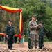 A military ceremony or briefing is taking place outdoors, featuring personnel in at least two distinct camouflage uniforms — one in US Marine Corps MARPAT digital camouflage and others in a darker woodland-pattern camouflage. The central figure, wearing USMC uniform and cover, is speaking at a microphone stand while holding a document. The setting is decorated with red and yellow draped fabric against a camouflage netting backdrop, suggesting a joint military exercise or formal event.