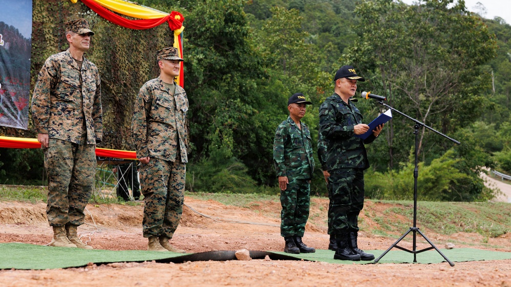 A military ceremony or briefing is taking place outdoors, featuring personnel from two different armed forces. Two individuals in US Marine Corps MARPAT digital camouflage uniforms stand at attention on the left, while two individuals in Thai military uniforms stand on the right, one of whom is speaking at a microphone podium. The setting appears to be a field environment with red clay ground, green matting, and a camouflage-decorated backdrop with red and yellow drapery.