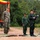 U.S. Marines and Thailand Mine Action Center participate in Deployed for Training Bilateral EOD Exercise A military ceremony or briefing is taking place outdoors, featuring personnel from two different armed forces. Two individuals in US Marine Corps MARPAT digital camouflage uniforms stand at attention on the left, while two individuals in Thai military uniforms stand on the right, one of whom is speaking at a microphone podium. The setting appears to be a field environment with red clay ground, green matting, and a camouflage-decorated backdrop with red and yellow drapery.
