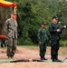 A military ceremony or briefing is taking place outdoors, featuring personnel from two different armed forces. Two individuals in US Marine Corps MARPAT digital camouflage uniforms stand at attention on the left, while two individuals in Thai military uniforms stand on the right, one of whom is speaking at a microphone podium. The setting appears to be a field environment with red clay ground, green matting, and a camouflage-decorated backdrop with red and yellow drapery.