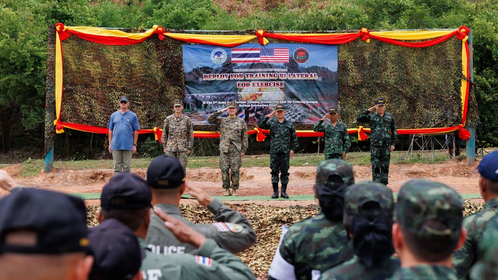 A formal military ceremony is taking place at the 'Deploy for Training Bi-Lateral EOD Exercise' held between August 14 and September 4, 2026, at the 2nd Army Area Tactical Training Ground (Phu Lam Yai Training Ground) in Nakhon Ratchasima Province, Thailand. The event involves both U.S. and Thai military personnel standing on a stage before an audience rendering salutes. The backdrop features Thai and American flags along with unit insignia, decorated with red and yellow bunting on camouflage netting.