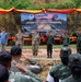 A formal military ceremony is taking place at the 'Deploy for Training Bi-Lateral EOD Exercise' held between August 14 and September 4, 2026, at the 2nd Army Area Tactical Training Ground (Phu Lam Yai Training Ground) in Nakhon Ratchasima Province, Thailand. The event involves both U.S. and Thai military personnel standing on a stage before an audience rendering salutes. The backdrop features Thai and American flags along with unit insignia, decorated with red and yellow bunting on camouflage netting.