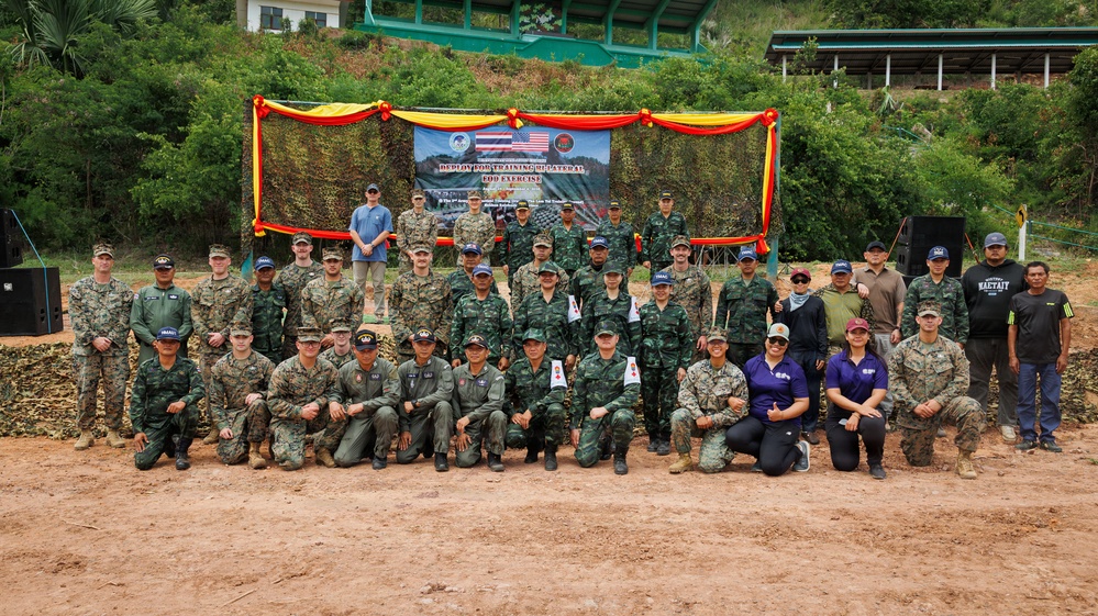A large group photo of military personnel from multiple nations posing together at what appears to be a joint military exercise or training event. A banner in the background references a 'Deployed Training Bilateral EOD Exercise,' with visible American and Thai flags suggesting a US-Thailand bilateral military cooperation event. Participants are wearing a mix of camouflage uniforms and civilian attire, arranged in three rows for the formal group photograph.