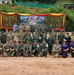 A large group photo of military personnel from multiple nations posing together at what appears to be a joint military exercise or training event. A banner in the background references a 'Deployed Training Bilateral EOD Exercise,' with visible American and Thai flags suggesting a US-Thailand bilateral military cooperation event. Participants are wearing a mix of camouflage uniforms and civilian attire, arranged in three rows for the formal group photograph.