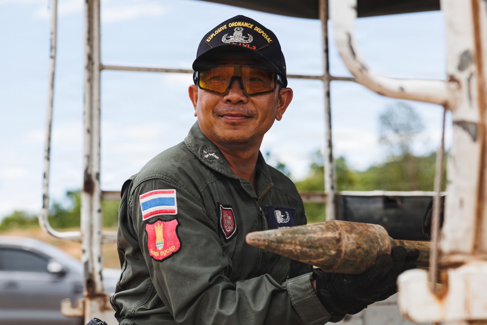 A Thai Explosive Ordnance Disposal (EOD) technician is pictured holding what appears to be an unidentified munition or ordnance item covered in dirt/mud. He is wearing a military flight suit with a Thai flag patch and a 'Bomb Squad' patch, along with an EOD cap and yellow-tinted safety glasses. The setting appears to be an outdoor field environment, likely during an EOD recovery or clearance operation.