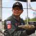 A Thai Explosive Ordnance Disposal (EOD) technician is pictured holding what appears to be an unidentified munition or ordnance item covered in dirt/mud. He is wearing a military flight suit with a Thai flag patch and a 'Bomb Squad' patch, along with an EOD cap and yellow-tinted safety glasses. The setting appears to be an outdoor field environment, likely during an EOD recovery or clearance operation.