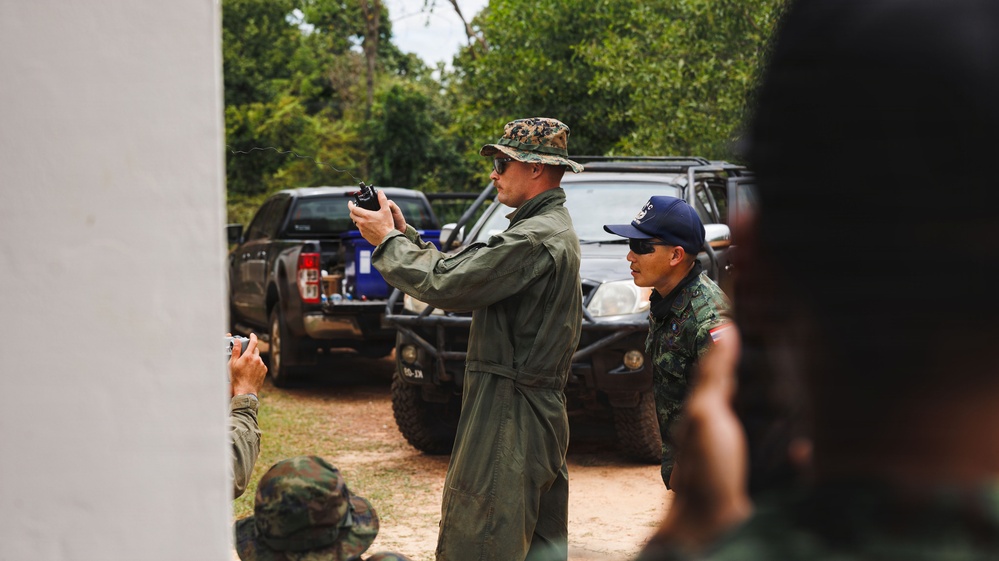 A military instructor in an olive green flight suit and camouflage boonie hat holds up a small device, appearing to conduct a demonstration or briefing for a group of soldiers. The setting is an outdoor training area with military and civilian vehicles parked in the background amid dense vegetation. Personnel in camouflage uniforms, including at least one individual wearing a blue cap with what appears to be a flag patch, are observing the instruction.