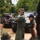 U.S. Marines and Thailand Mine Action Center participate in Deployed for Training Bilateral EOD Exercise A military instructor in an olive green flight suit and camouflage boonie hat holds up a small device, appearing to conduct a demonstration or briefing for a group of soldiers. The setting is an outdoor training area with military and civilian vehicles parked in the background amid dense vegetation. Personnel in camouflage uniforms, including at least one individual wearing a blue cap with what appears to be a flag patch, are observing the instruction.