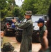 A military instructor in an olive green flight suit and camouflage boonie hat holds up a small device, appearing to conduct a demonstration or briefing for a group of soldiers. The setting is an outdoor training area with military and civilian vehicles parked in the background amid dense vegetation. Personnel in camouflage uniforms, including at least one individual wearing a blue cap with what appears to be a flag patch, are observing the instruction.