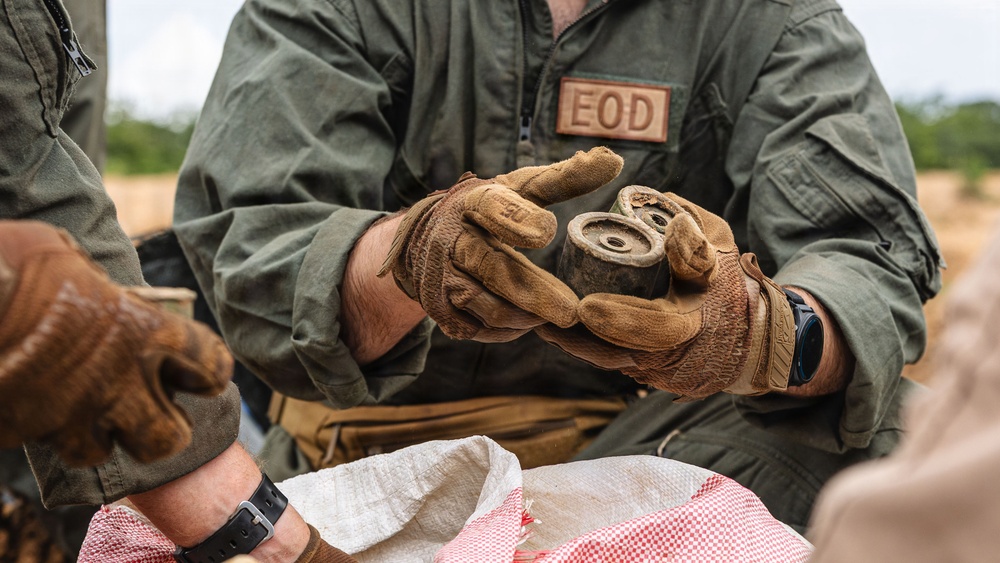 An EOD (Explosive Ordnance Disposal) technician wearing a green flight suit and tan tactical gloves is handling what appears to be a small munition or ordnance component. The individual is working in an outdoor field environment alongside at least one other person. A woven sack with a red-and-white pattern is visible in the foreground.
