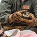 An EOD (Explosive Ordnance Disposal) technician wearing a green flight suit and tan tactical gloves is handling what appears to be a small munition or ordnance component. The individual is working in an outdoor field environment alongside at least one other person. A woven sack with a red-and-white pattern is visible in the foreground.