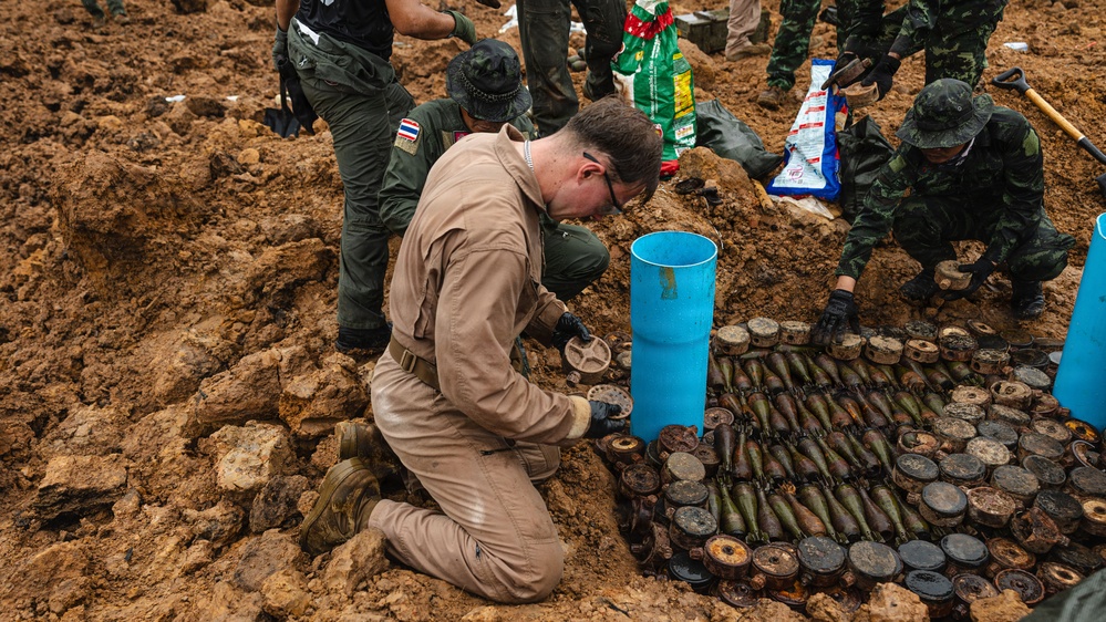 The image shows what appears to be an explosive ordnance disposal (EOD) operation, likely in Southeast Asia based on the Thai military uniforms visible. A specialist in tan/khaki coveralls is kneeling over a large cache of unearthed munitions, including what appear to be mortar rounds, landmines, and other ordnance laid out in rows. Military personnel in camouflage uniforms are working in the background at an excavation site.