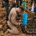The image shows what appears to be an explosive ordnance disposal (EOD) operation, likely in Southeast Asia based on the Thai military uniforms visible. A specialist in tan/khaki coveralls is kneeling over a large cache of unearthed munitions, including what appear to be mortar rounds, landmines, and other ordnance laid out in rows. Military personnel in camouflage uniforms are working in the background at an excavation site.