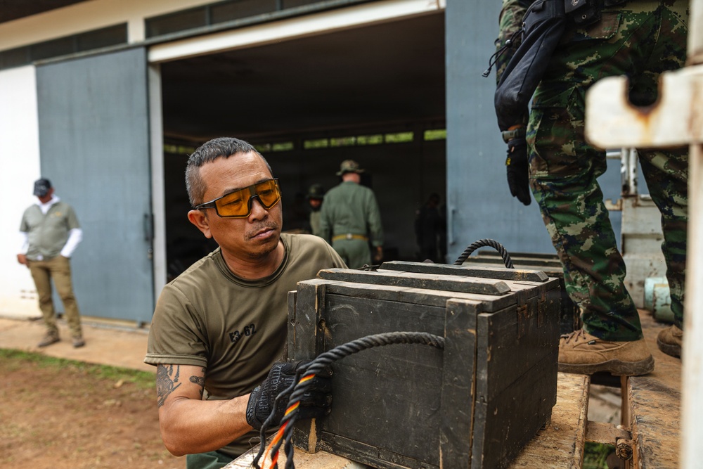 A man wearing amber safety glasses, an olive green military-style t-shirt with markings 'E3-02', and tactical gloves is handling a heavy dark wooden or metal crate on what appears to be a vehicle or platform. Several other individuals in military/tactical uniforms are visible in the background near an open hangar or warehouse structure. The setting appears to be a military or law enforcement operational environment.