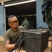 A man wearing amber safety glasses, an olive green military-style t-shirt with markings 'E3-02', and tactical gloves is handling a heavy dark wooden or metal crate on what appears to be a vehicle or platform. Several other individuals in military/tactical uniforms are visible in the background near an open hangar or warehouse structure. The setting appears to be a military or law enforcement operational environment.