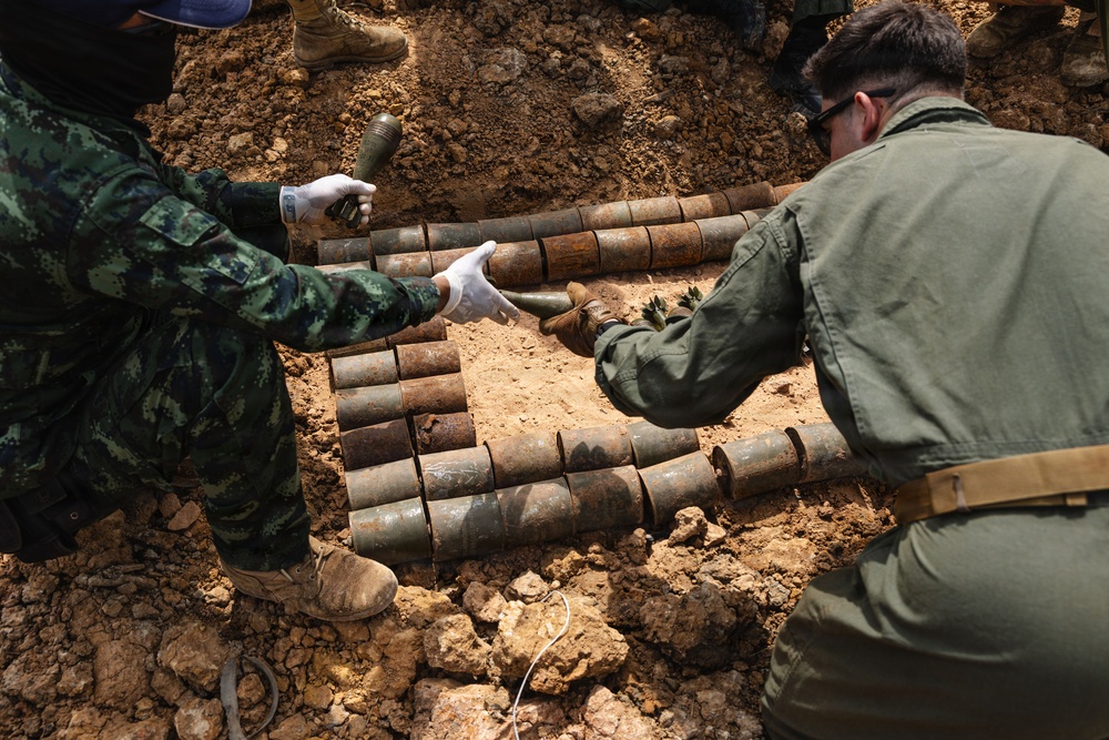 Two personnel in military/tactical clothing are handling a large collection of corroded, cylindrical munitions or ordnance items that have been excavated from the ground. The items appear to be old, heavily rusted artillery shells or similar explosive ordnance arranged in rows on disturbed soil. This appears to be an unexploded ordnance (UXO) clearance or explosive ordnance disposal (EOD) operation.