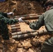 Two personnel in military/tactical clothing are handling a large collection of corroded, cylindrical munitions or ordnance items that have been excavated from the ground. The items appear to be old, heavily rusted artillery shells or similar explosive ordnance arranged in rows on disturbed soil. This appears to be an unexploded ordnance (UXO) clearance or explosive ordnance disposal (EOD) operation.