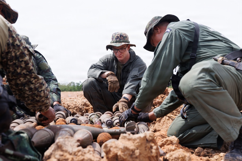 Multiple military or security personnel in different uniforms are crouched over a collection of what appear to be recovered ordnance or munitions partially buried in sandy soil. A central figure wearing a camouflage boonie hat, glasses, and tactical gloves is closely examining the items. The scene suggests a demining, explosive ordnance disposal (EOD), or weapons cache documentation operation.