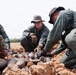 Multiple military or security personnel in different uniforms are crouched over a collection of what appear to be recovered ordnance or munitions partially buried in sandy soil. A central figure wearing a camouflage boonie hat, glasses, and tactical gloves is closely examining the items. The scene suggests a demining, explosive ordnance disposal (EOD), or weapons cache documentation operation.