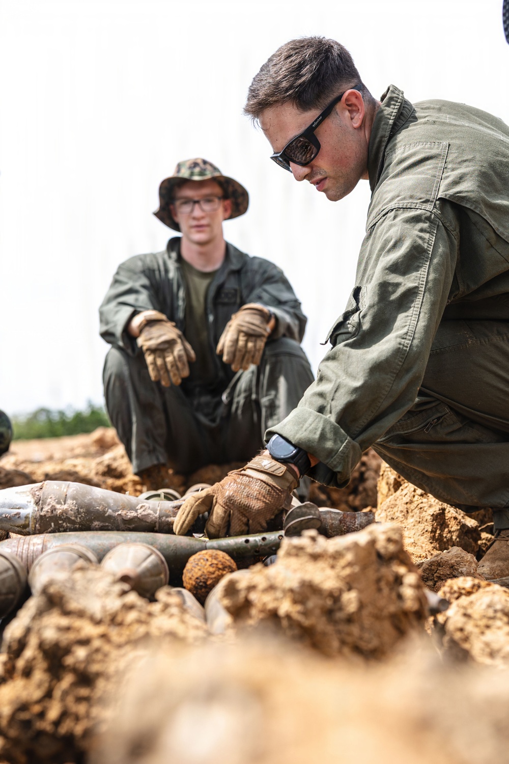 Two individuals in olive/green military-style uniforms and tactical gloves are examining a collection of what appear to be excavated munitions or ordnance items partially buried in sandy soil. The person in the foreground is leaning down and handling the items, while the second person in the background observes while seated, wearing a camouflage boonie hat and glasses. The scene is consistent with an Explosive Ordnance Disposal (EOD) or battlefield archaeology/clearance operation.