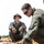 U.S. Marines and Thailand Mine Action Center participate in Deployed for Training Bilateral EOD Exercise Two individuals in olive/green military-style uniforms and tactical gloves are examining a collection of what appear to be excavated munitions or ordnance items partially buried in sandy soil. The person in the foreground is leaning down and handling the items, while the second person in the background observes while seated, wearing a camouflage boonie hat and glasses. The scene is consistent with an Explosive Ordnance Disposal (EOD) or battlefield archaeology/clearance operation.