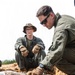 Two individuals in olive/green military-style uniforms and tactical gloves are examining a collection of what appear to be excavated munitions or ordnance items partially buried in sandy soil. The person in the foreground is leaning down and handling the items, while the second person in the background observes while seated, wearing a camouflage boonie hat and glasses. The scene is consistent with an Explosive Ordnance Disposal (EOD) or battlefield archaeology/clearance operation.