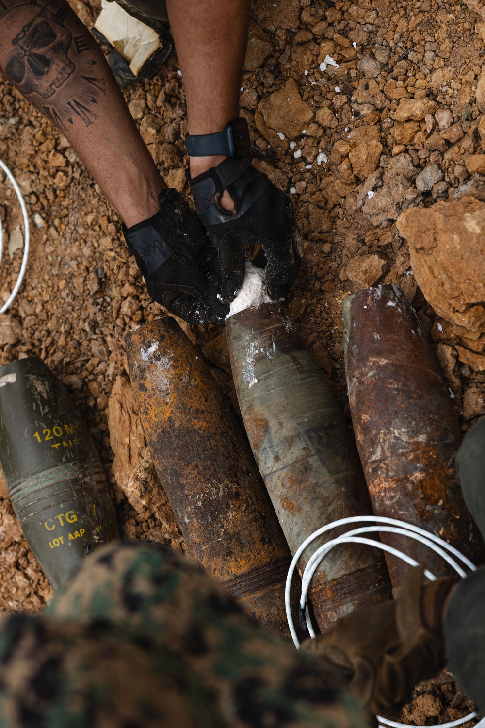 A person wearing black tactical gloves and camouflage uniform is handling multiple large corroded mortar rounds or artillery shells in an excavated dirt pit. One visible shell is marked '120MM TNT' and 'CTG LOT AAP', indicating it is a 120mm mortar cartridge. White detonation cord (det cord) is visible near the ordnance, suggesting an Explosive Ordnance Disposal (EOD) or demolition operation.