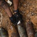 A person wearing black tactical gloves and camouflage uniform is handling multiple large corroded mortar rounds or artillery shells in an excavated dirt pit. One visible shell is marked '120MM TNT' and 'CTG LOT AAP', indicating it is a 120mm mortar cartridge. White detonation cord (det cord) is visible near the ordnance, suggesting an Explosive Ordnance Disposal (EOD) or demolition operation.