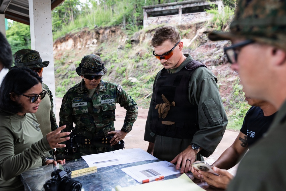 A group of military personnel and a civilian woman are gathered around a table reviewing documents and maps in an outdoor covered area. The group appears to include both Thai military soldiers in camouflage uniforms and Western military personnel in tactical gear. The setting suggests a joint military planning or briefing session.