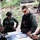 U.S. Marines and Thailand Mine Action Center participate in Deployed for Training Bilateral EOD Exercise A group of military personnel and a civilian woman are gathered around a table reviewing documents and maps in an outdoor covered area. The group appears to include both Thai military soldiers in camouflage uniforms and Western military personnel in tactical gear. The setting suggests a joint military planning or briefing session.