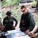 A group of military personnel and a civilian woman are gathered around a table reviewing documents and maps in an outdoor covered area. The group appears to include both Thai military soldiers in camouflage uniforms and Western military personnel in tactical gear. The setting suggests a joint military planning or briefing session.