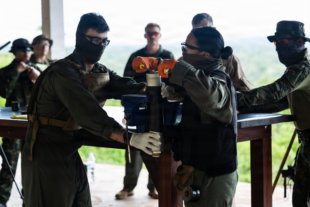 Military or law enforcement personnel in tactical gear are working together at a workbench, manipulating what appears to be a bench vise or similar tool-holding device. The individuals are wearing protective equipment including face coverings, safety glasses, gloves, and body armor. The setting appears to be an outdoor or semi-open training facility with vegetation visible in the background.
