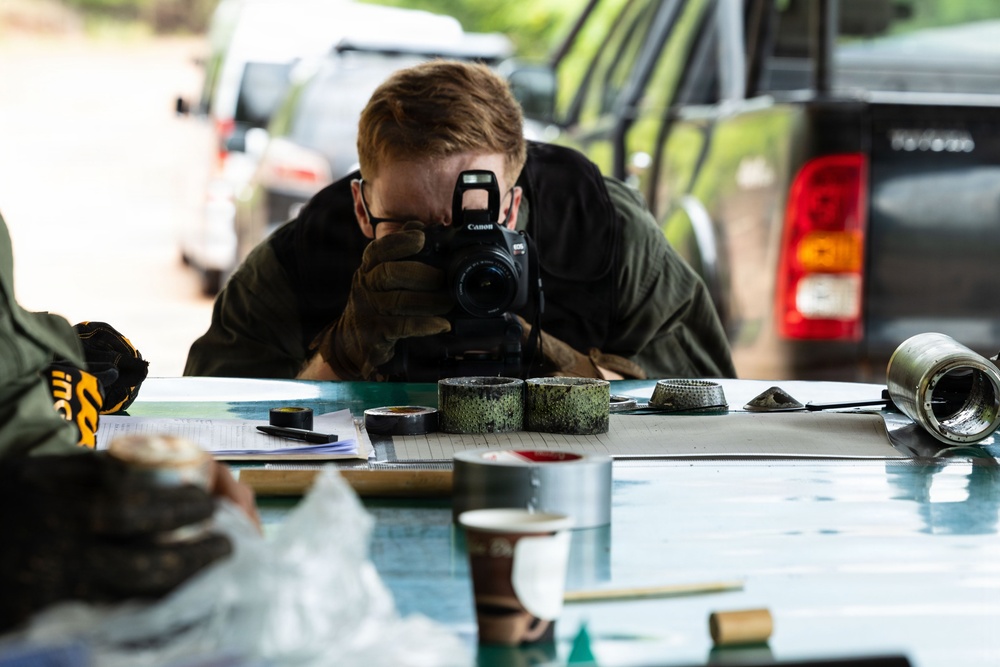 A person wearing gloves and a dark jacket is crouched low over a table, photographing items with a Canon DSLR camera. The table contains various objects including rolls of tape, documents, containers, and cylindrical metal objects. The setting appears to be an outdoor or semi-covered area with vehicles visible in the background.