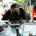 A person wearing gloves and a dark jacket is crouched low over a table, photographing items with a Canon DSLR camera. The table contains various objects including rolls of tape, documents, containers, and cylindrical metal objects. The setting appears to be an outdoor or semi-covered area with vehicles visible in the background.