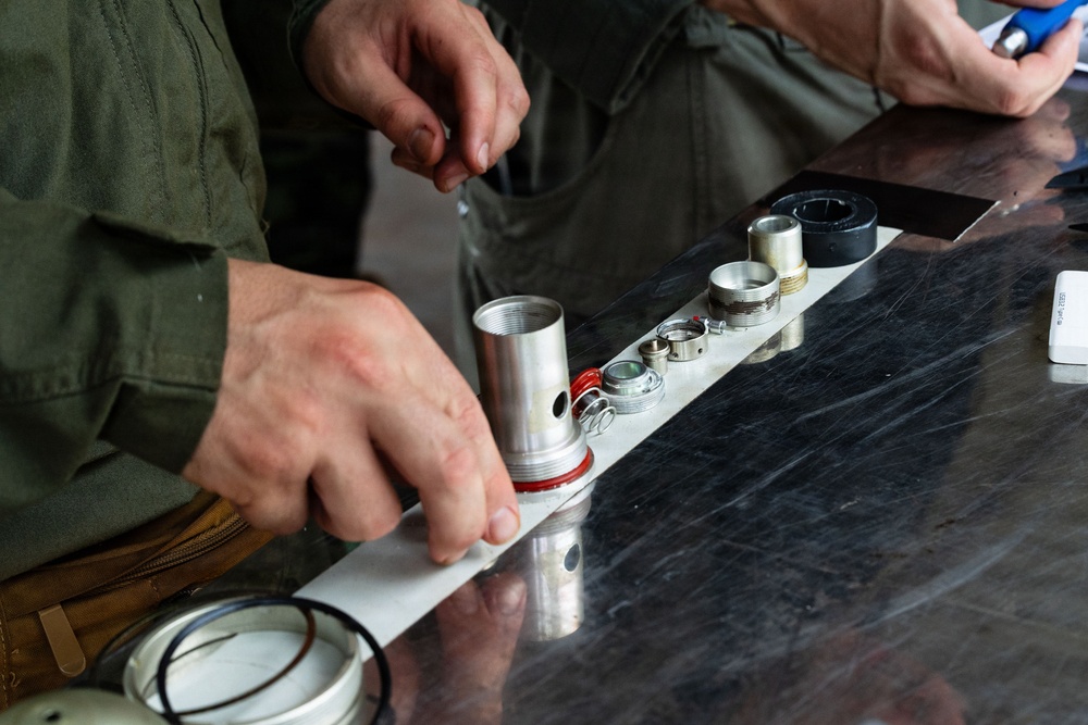 A person wearing an olive/green work shirt is assembling or disassembling mechanical components on a dark marble or stone surface. Multiple cylindrical metal parts, rings, and fittings are laid out in a row on a white strip or rail. A second person is partially visible in the background, appearing to observe.