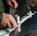 A person wearing an olive/green work shirt is assembling or disassembling mechanical components on a dark marble or stone surface. Multiple cylindrical metal parts, rings, and fittings are laid out in a row on a white strip or rail. A second person is partially visible in the background, appearing to observe.