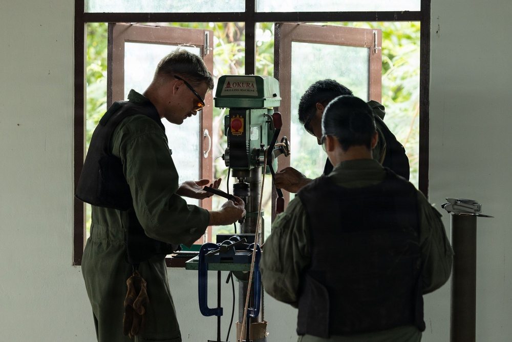 Three individuals wearing olive/dark green uniforms and protective eyewear are gathered around an OKURA brand drill press machine in what appears to be a workshop or training facility. They appear to be operating or inspecting the drilling machine together. Natural vegetation is visible through the windows behind them.