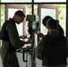 Three individuals wearing olive/dark green uniforms and protective eyewear are gathered around an OKURA brand drill press machine in what appears to be a workshop or training facility. They appear to be operating or inspecting the drilling machine together. Natural vegetation is visible through the windows behind them.