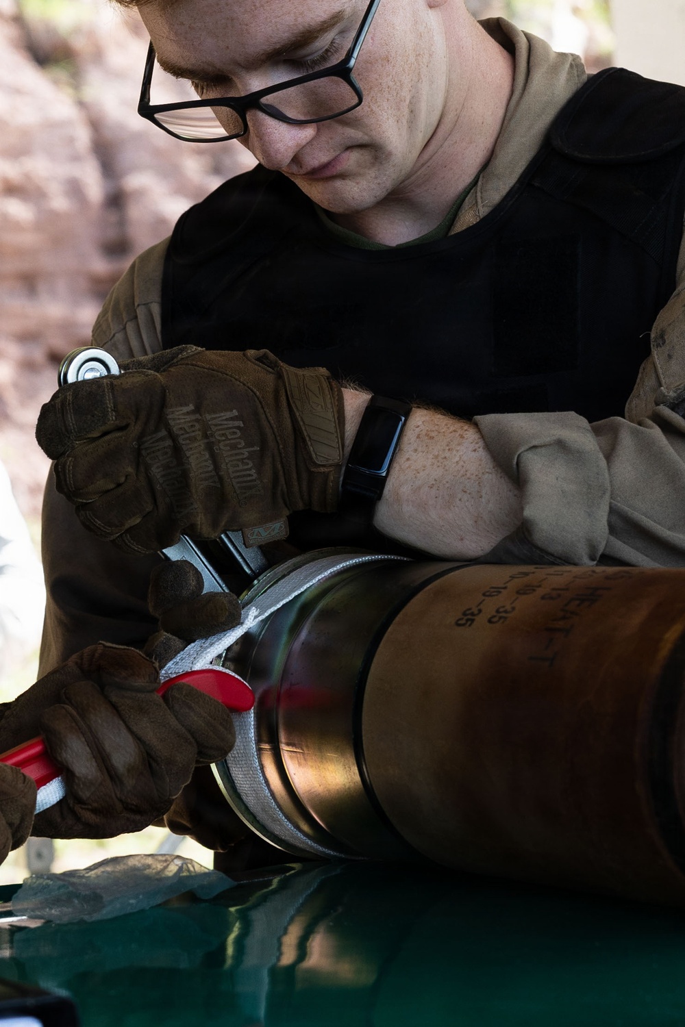 A young male technician wearing glasses and Mechanix brand tactical gloves is working on a large cylindrical metallic object, using red-handled pliers or cutters to remove or cut a fabric/tape band. The individual is dressed in military-style olive/tan clothing and a black undershirt, working outdoors in what appears to be a field environment. Stenciled markings are visible on the cylindrical object.
